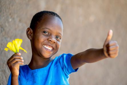 Ein kleines Mädchen lächelt. Es streckt den Daumen der einen Hand hoch, in der anderen Hand hält es eine gelbe Blüte.