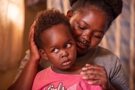 Eine Frau legt die Hände auf Kopf und Schulter eines kleinen Mädchen, das die Augen nach oben dreht und weiße Pupillen hat.