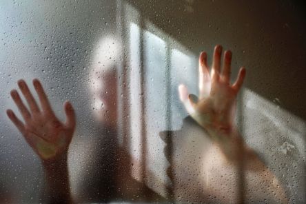 Zwei Hände auf einer nassen Glasscheibe, dahinter die Silhouette einer Frau. Auf der Scheibe spiegelt sich ein Fenster. Ein Lichtschein wirft den Schatten der Frau auf die Scheibe. 