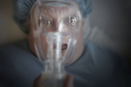 Eine Frau mit aufgrissenen Augen und offenem Mund in OP-Kleidung schaut durch eine Narkosemaske vor ihrem Gesicht.