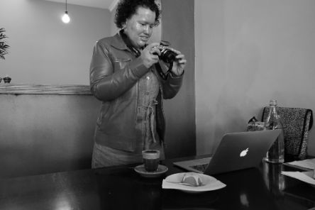 Eine Frau steht hinter einem Tisch auf dem ein Laptop, Kaffee und Kuchen steht. Sie hält eine Fotokamera in der Hand und blickt auf diese.