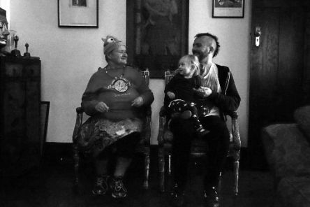 Eine Frau und ein Mann sitzen nebeneinander auf Stühlen, schauen sich an und lachen. Der Mann hat ein Kind auf dem Schoß.