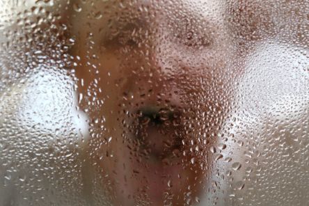 Eine Frau steht frontal hinter einer Glasscheibe voller Wassertropfen. Sie hält sich mit beiden Händen den Kopf und schreit mit weit geöffnetem Mund. 