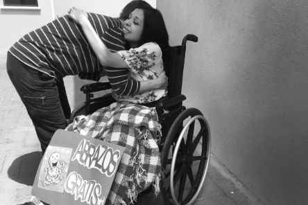 Eine Frau im Rollstuhl und eine Person, die vor ihr steht, umarmen sich. Vor der Frau im Rollstuhl steht ein Schild mit "Abrazos Gratis", was "kostenlose Umarmungen" bedeutet.