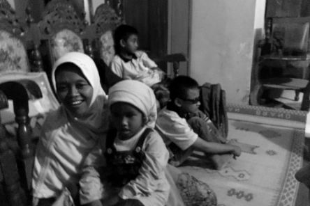 Eine lachende Frau mit Kopftuch sitzt vor Polsterstühlen auf einem Webteppich mit einem Kind auf dem Schoß, im Hintergrund zwei Jungen.