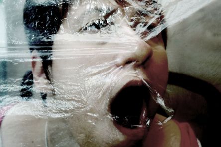 Frauengesicht hinter einer Folie. Es ist unklar, ob sie ihr Gesicht leicht in die Folie drückt oder ob die Folie auf ihr Gesicht gedrückt wird.