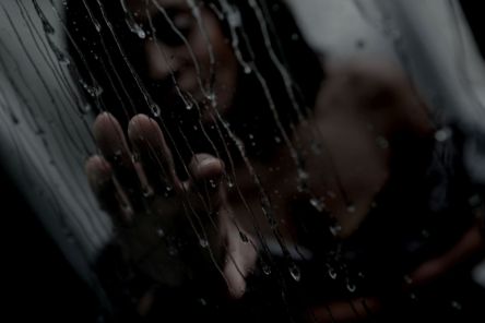 Eine Frau hinter einer Scheibe voller Wassertropfen hat ihre Hand auf die Scheibe gelet.