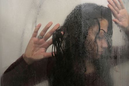 Eine Frau mit langem, dunklem Haar legt ihre Hände auf eine nasse Scheibe, hinter der sie steht.