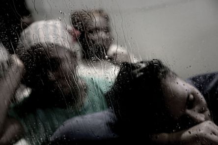 Drei Frauen sitzen hintereinander. Die Vorderste legt ihren Kopf auf ihren Arm ab, die zweite stützt ihren Kopf auf der rechten Hand auf. Dahinter ist das Gesicht einer dritten Frau zu erkennen. Ihre Gesichter sind unscharf, sie befinden sich hinter einer nassen Glasscheibe.