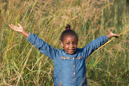 Ein in einer Wiese stehendes afrikanisches Mädchen hält beide Arme hoch.