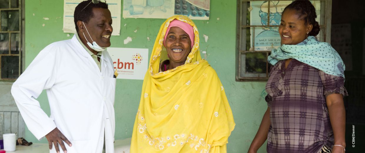 Mann im weißen Kittel, lachende Frau im gelben Gewand und eine weitere Frau, die Papiere in der Hand hält.