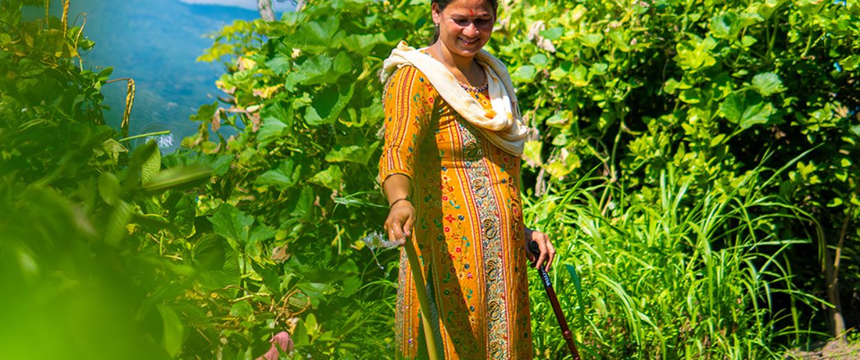Eine etwa 30-jährige Frau steht mit Gehstock auf einem Feld. In der rechten Hand hält sie einen Gartenschlauch, aus dem Wasser spritzt.