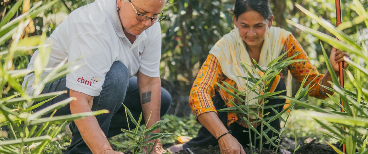 Eine Frau im Sari mit Stock und eine Frau in Jeans und Polohemd hocken auf einem Feld und klopfen mit den Händen die Erde um kleine Grünpflanzen fest.