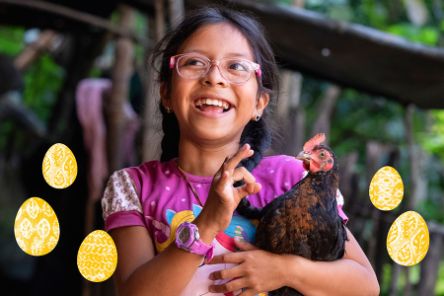 Ein etwa zehnjähriges Mädchen mit geflochtenen Zöpfen hält lachend ein kleines Huhn im Arm.