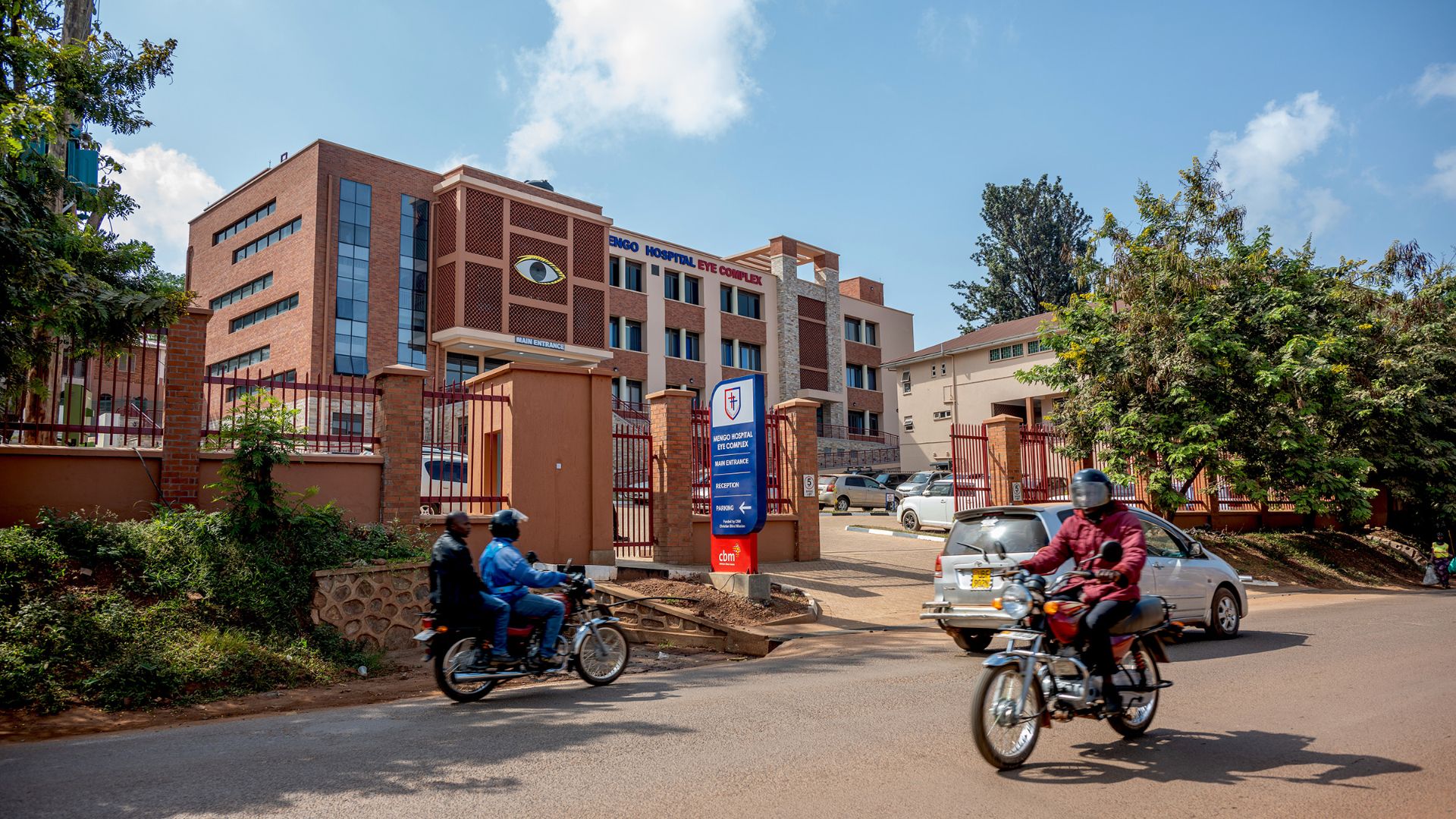 Ein Transporter mit der Aufschrift &quot;Mengo Hospital&quot; fährt aus dem umzäunten Parkplatz vor einem mehrgeschossigen Gebäudekomplex.
