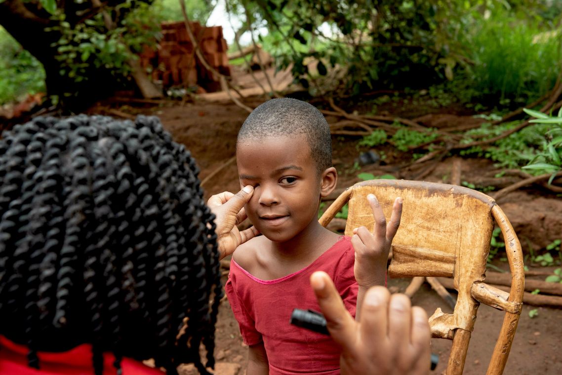 Eine Frau hat einen Finger auf das Augenlid eines Kindes gelegt und hält den rechten Zeigefinger hoch. Das Kind hält zwei Finger hoch.