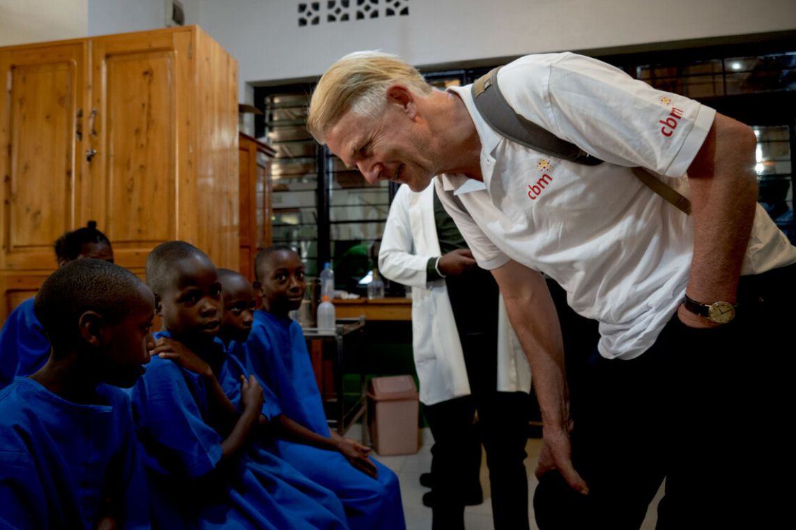 Ein Mann beugt sich zu den Kindern, die auf einer Wartebank sitzen. Sie tragen blaue Hemden.