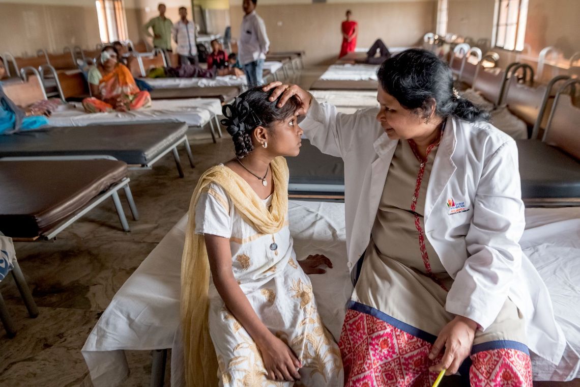 Eine Frau im weißen Kittel sitzt neben einem Mädchen auf einem Krankenbett und untersucht seine Augen.