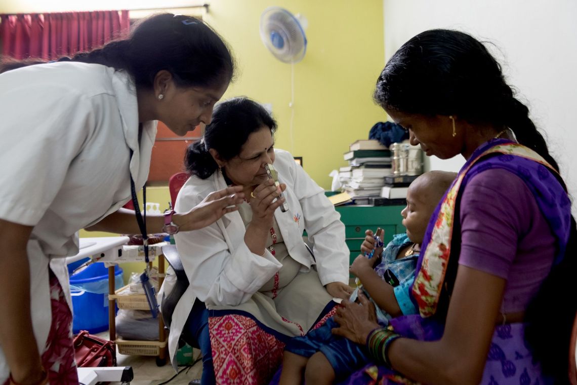 Eine Frau im weißen Kittel hält einem Kleinkind ein Handydisplay hin. Das Kind sitzt auf dem Schoß einer Frau.