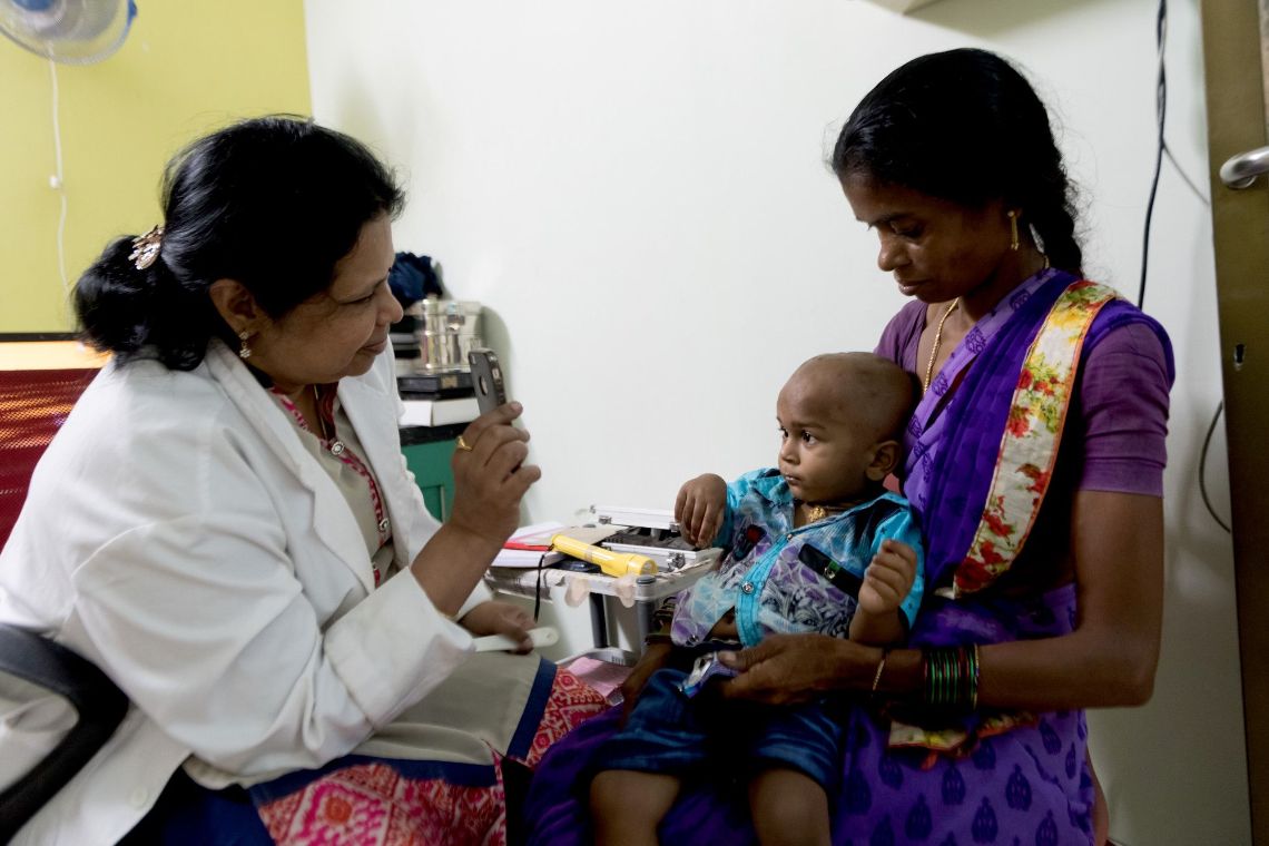 Eine Frau im weißen Kittel hält einem Kleinkind das Handy hin. Das Kind sitzt auf dem Schoß einer Frau im Sari.