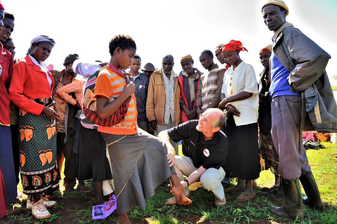Ein Mann untersucht den Fuß einer Frau, eine Gruppe Menschen steht im Halbkreis darum herum.