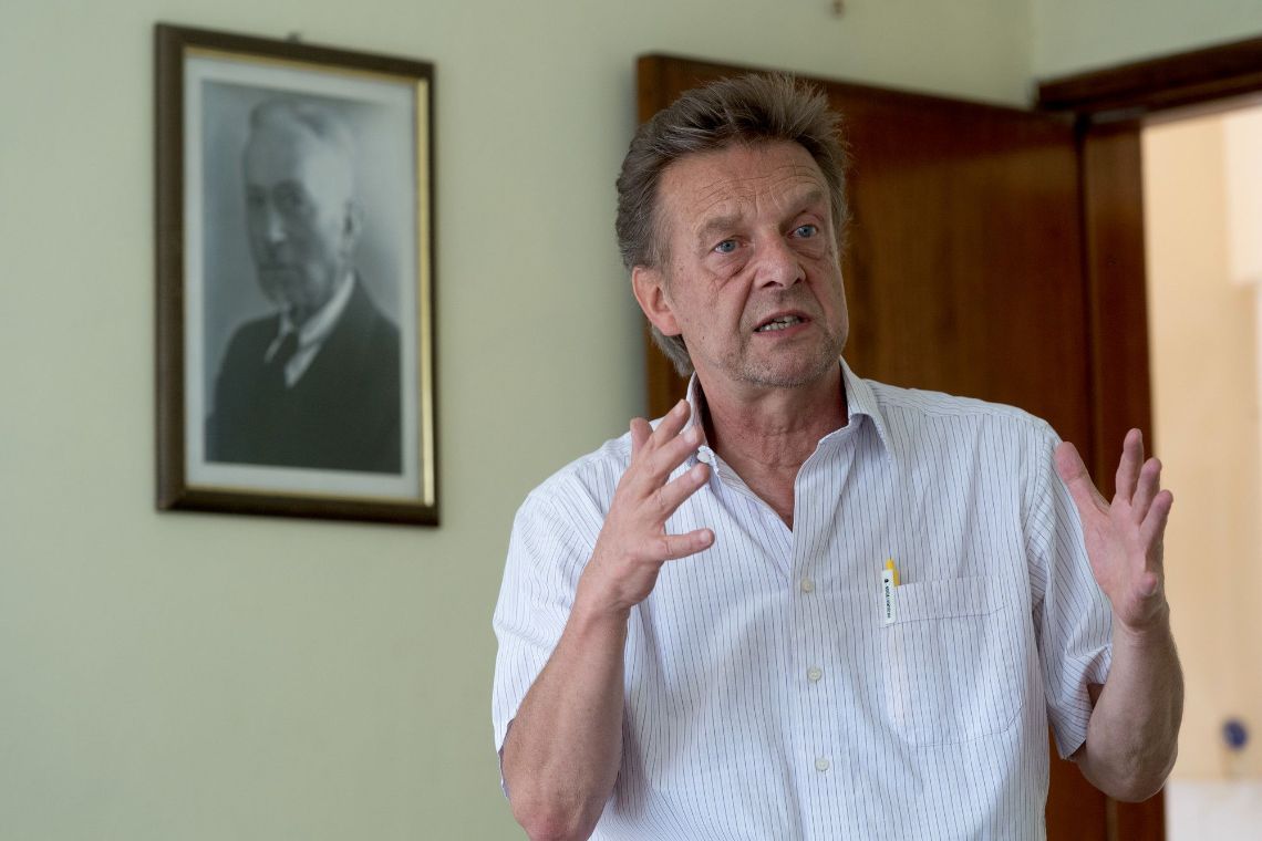 Ein Mann spricht und macht dabei Gesten mit den Händen, an der Wand hinter ihm hängt das Fot eines Mannes im Anzug.