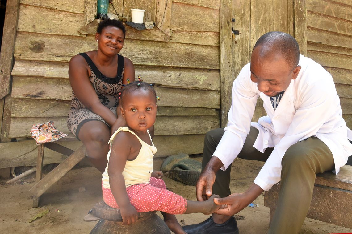 Ein Mann im weißen Kittel untersucht den Fuß eines kleinen Mädchens, eine Frau schaut zu.