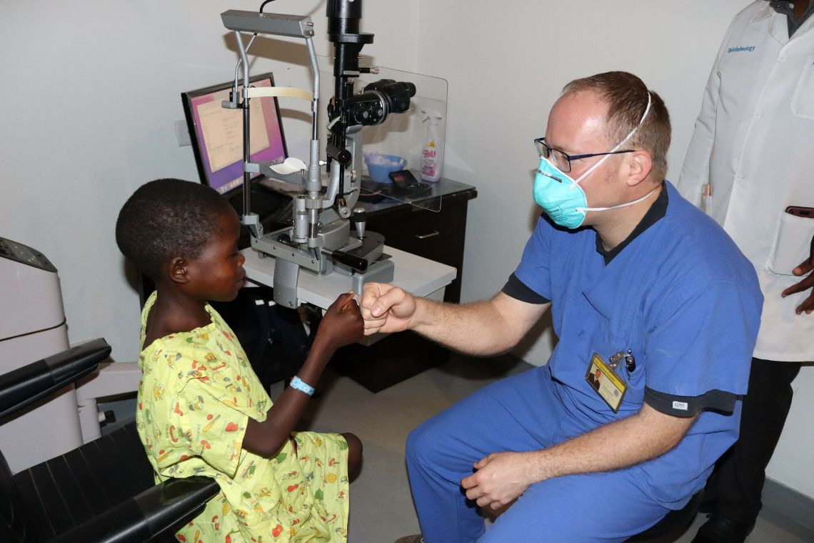 Ein Mann in OP-Kleidung mit Mund-Nasen-Schutz und ein kleines Mädchen begrüßen sich mit der Berührung der Fäuste.