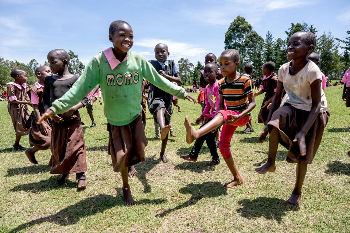 Ein Mädchen hüpft lachend zwischen anderen Kindern auf einer Wiese.