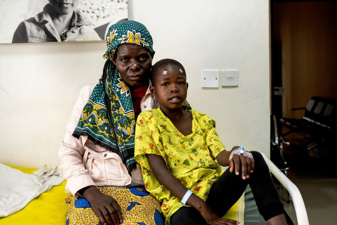 Eine Frau und ein Mädchen sitzen auf einem Krankenhausbett. Das Kind hat Markierungspflastern auf der Stirn über beiden Augen.