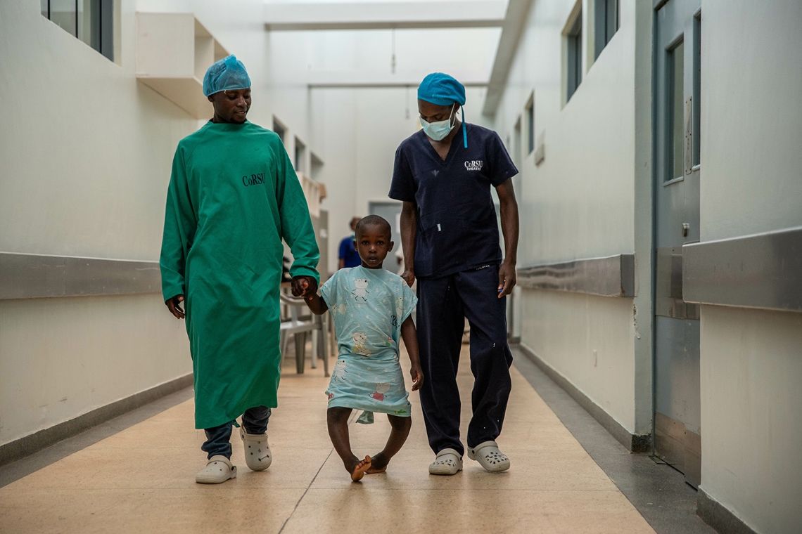 Ein kleiner Junge mit O-Beinen in OP-Kleidung wird von zwei Männern an der Hand geführt, sie gehen einen Flur entlang.