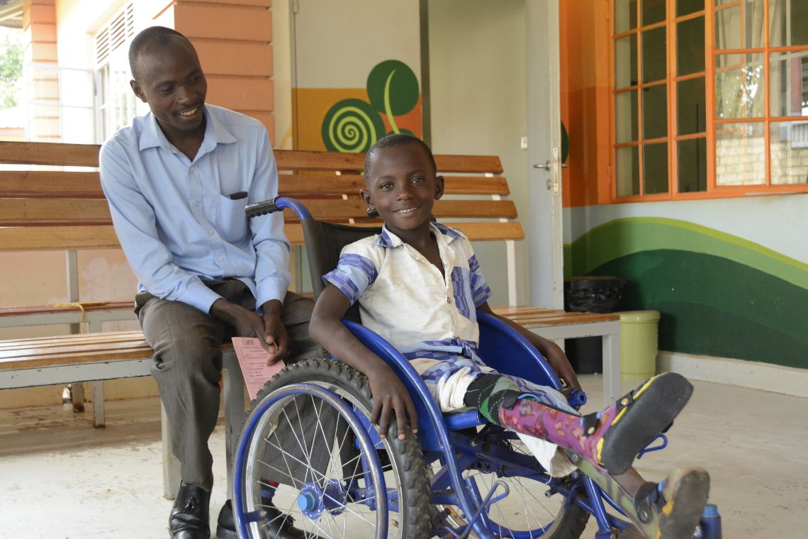 Ein lachender Junge im Rollstuhl; hinter dem Rollstuhl sitzt ein Mann auf einer Holzbank.