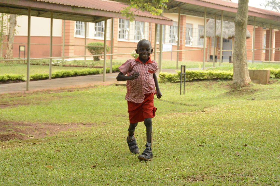 Ein kleiner Junge mit Beinschienen rennt auf einer Wiese, im Hintergrund ein eingschossiges Gebäude