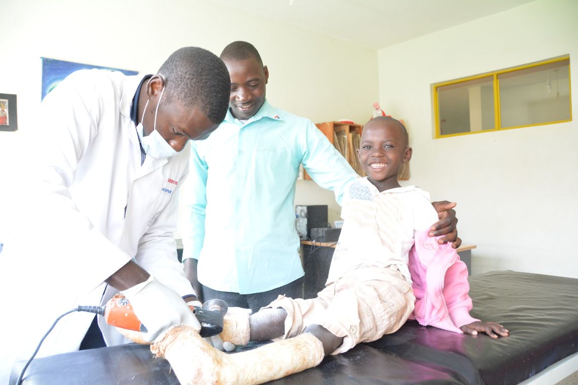 Ein Mann sägt den Gipsverband eines Kindes auf. Ein anderer Mann legt seine Hand auf die Schulter des Jungen.