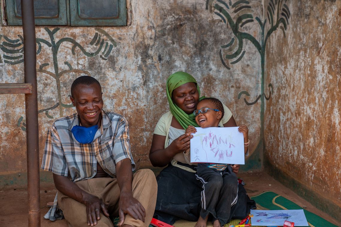 Lachende Menschen – ein Mann, eine Frau, ein kleines Mädchen – auf der Veranda eines einfachen Häuschens. Das Kind hält ein Blatt in der Hand, auf dem &quot;Thank you&quot; steht.