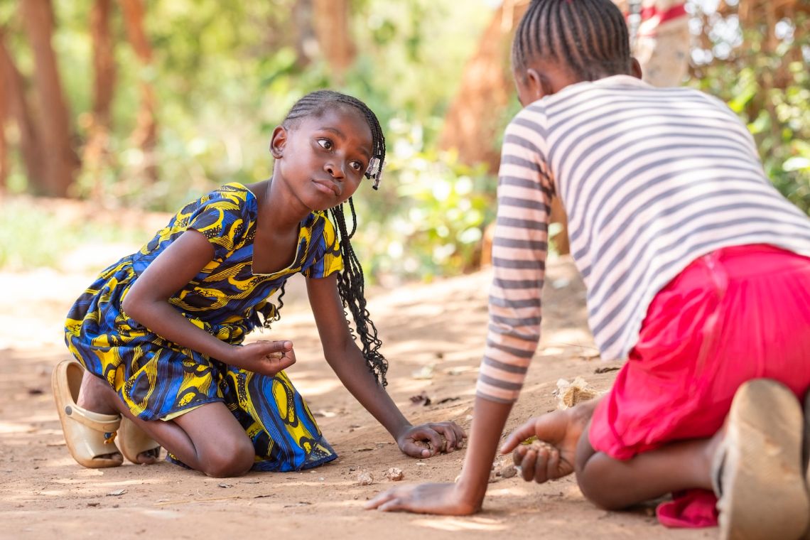 Ein etwa Neunjährige und etwa Zwölfjährige knieen am Boden und spielen mit Steinchen.