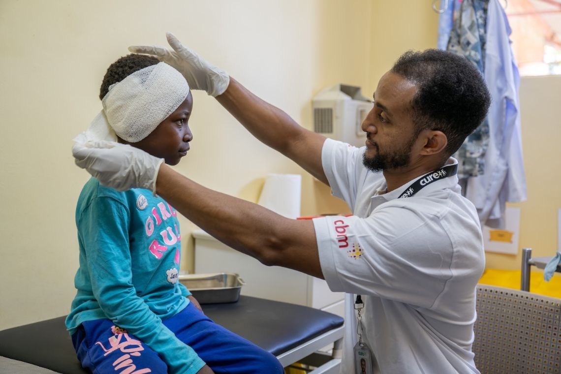 Ein Mann nimmt einem etwa zehnjährigen Mädchen den Ohrenverband ab.