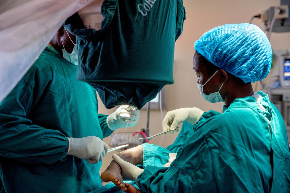 Ein kleines afrikanisches Mädchen mit extremen O-Beinen lehnt sich an einen Baumstamm.