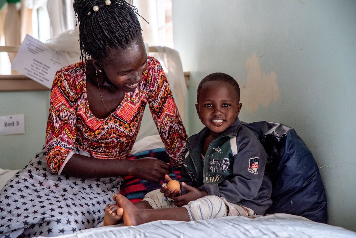 Ein kleines afrikanisches Mädchen sitzt auf einem Bett und lacht; neben ihm eine Frau.