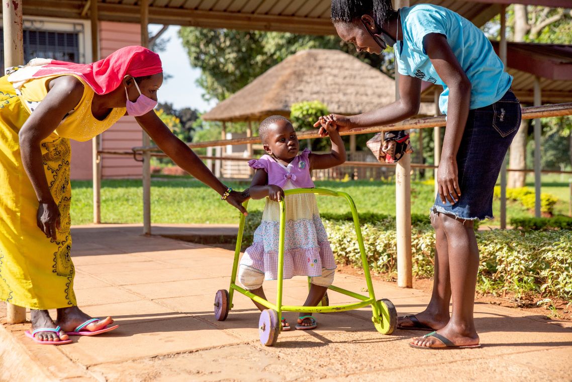 Ein kleines afrikanisches Mädchen geht an einem Rollator, begleitet von zwei Frauen.