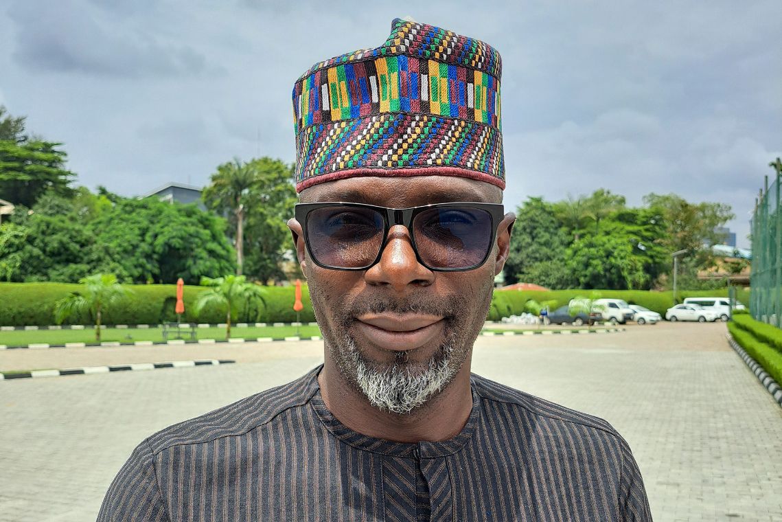 Ein Mann mit bunter Kappe und großer Sonnenbrille steht auf einem gepflasterten Platz, im Hintergrund sind Autos geparkt.