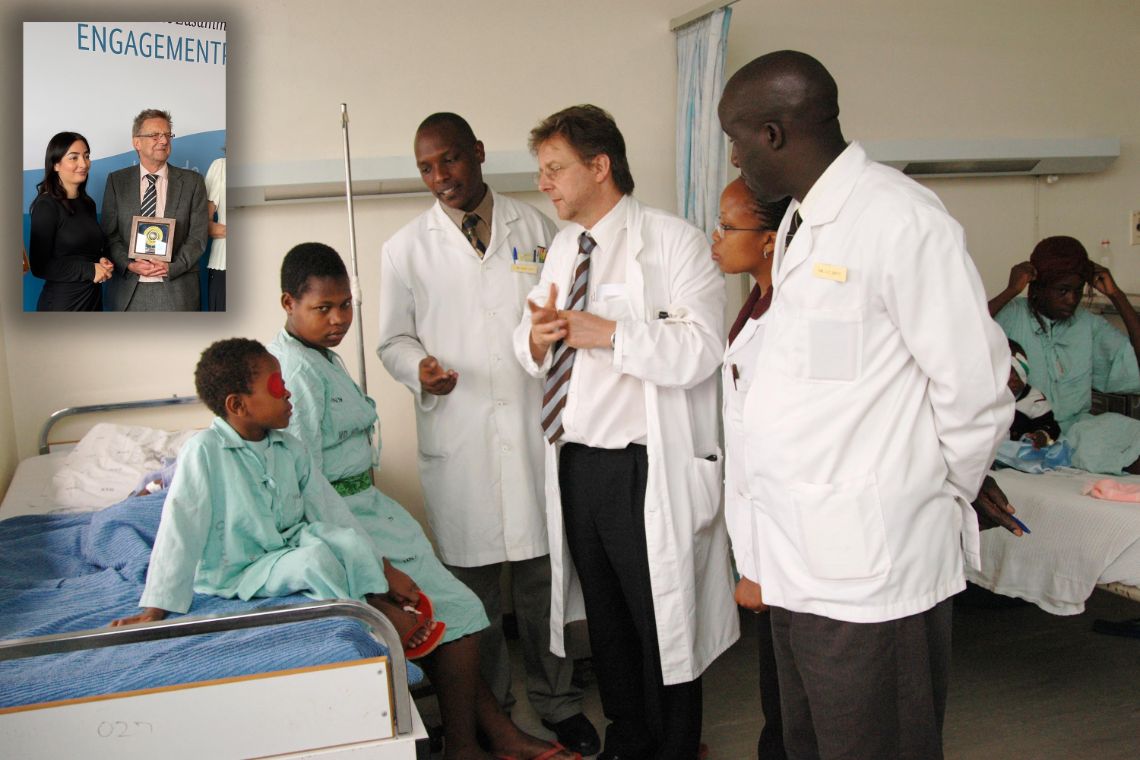 Männer und Frauen in weißen Kitteln stehen vor einem Krankenbett, auf dem ein Kind und eine weitere Person sitzen. Beide tragen ein grünes Nachthemd.