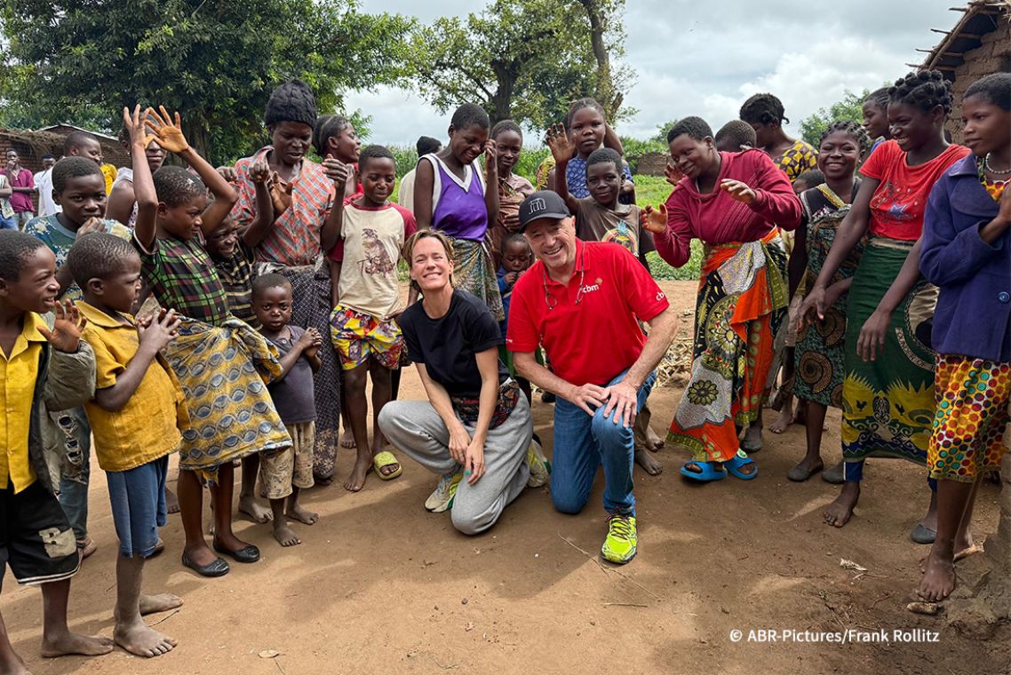 Eine Gruppe von Kindern und Frauen umringt einen älteren Mann und eine etwa vierzigjährige Frau, die halb in der Hocke, halb knieend am Boden sitzen. Die meisten lachen oder lächeln.