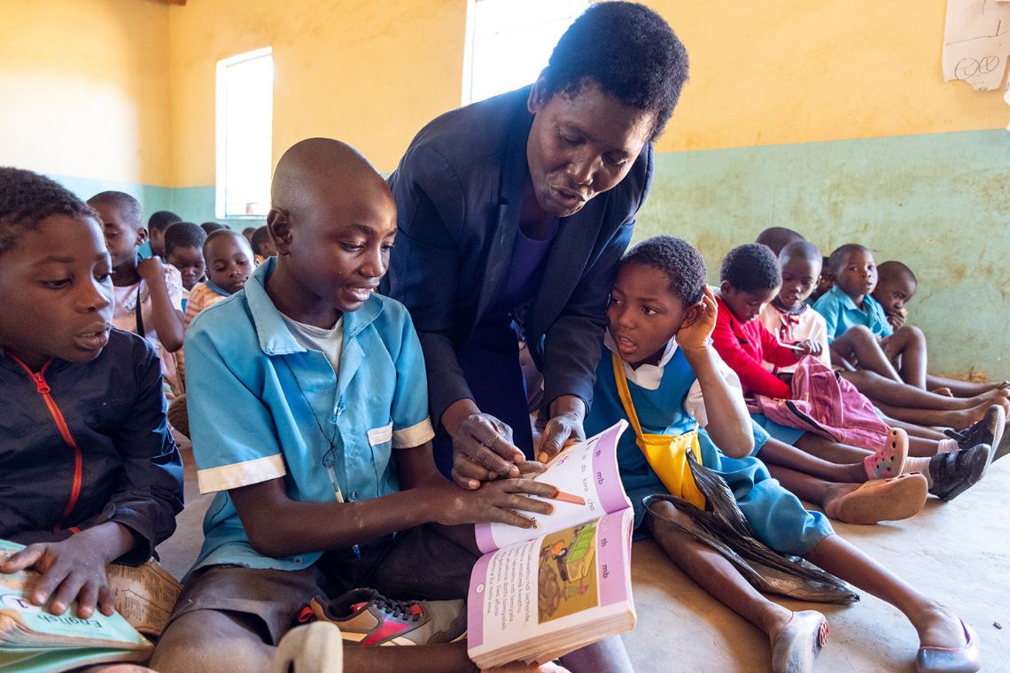 Eine Frau beugt sich in einem Klassenzimmer über die Schulter eines Jungen und zeigt auf die offene Seite des Heftes, das der Junge in der Hand hält.