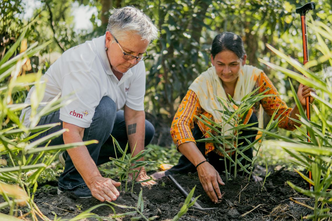 Eine Frau im Sari und eine Frau mit CBM-Polohemd sitzen in einem Beet mit noch kleinen Pfanzen.