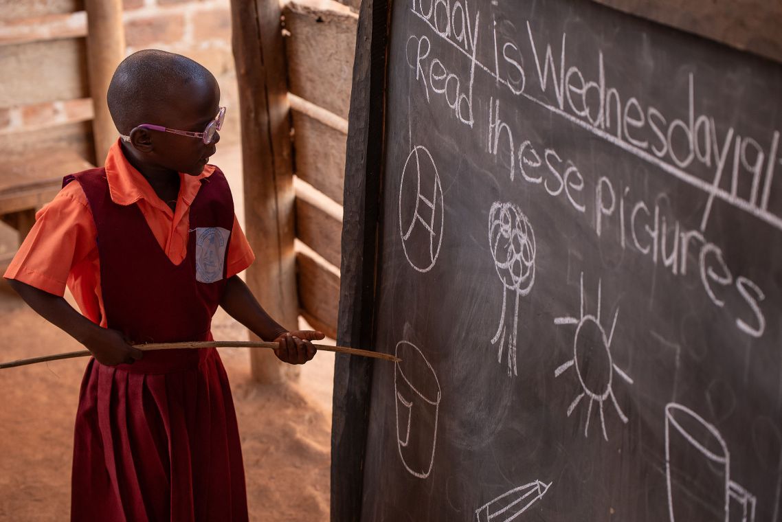 Mädchen in Schuluniform steht mit einem Zeigestock vor einer Tafel.