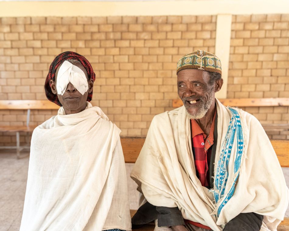 Eine Frau mit Augenverbänden sitzt neben ihrem Ehemann auf einer Bank.