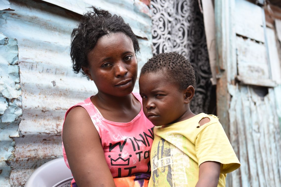Eine Frau mit einem etwa siebenjährigen Jungen auf dem Schoß sitzt vor einer Hütte.