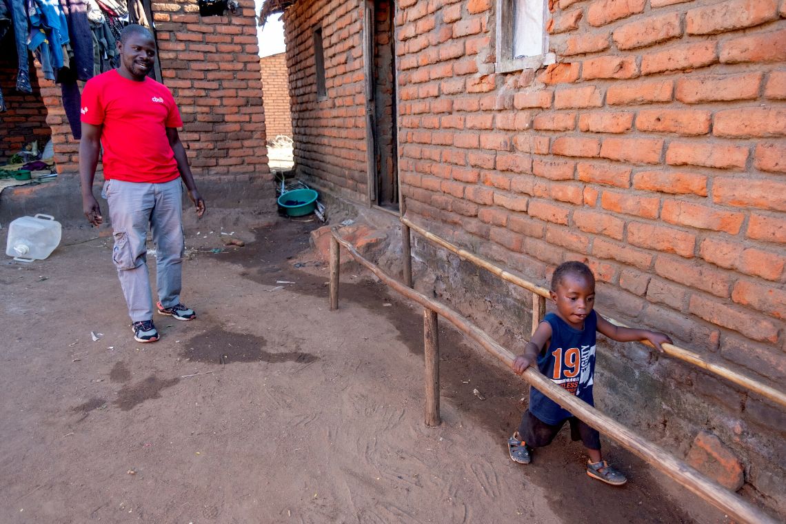 Ein kleiner Junge läuft an einem einfachen Holzbarren, an dem er sich mit beiden Händen festhält. Ein Mann im CBM-T-Shirt schaut ihm lächelnd dabei zu.