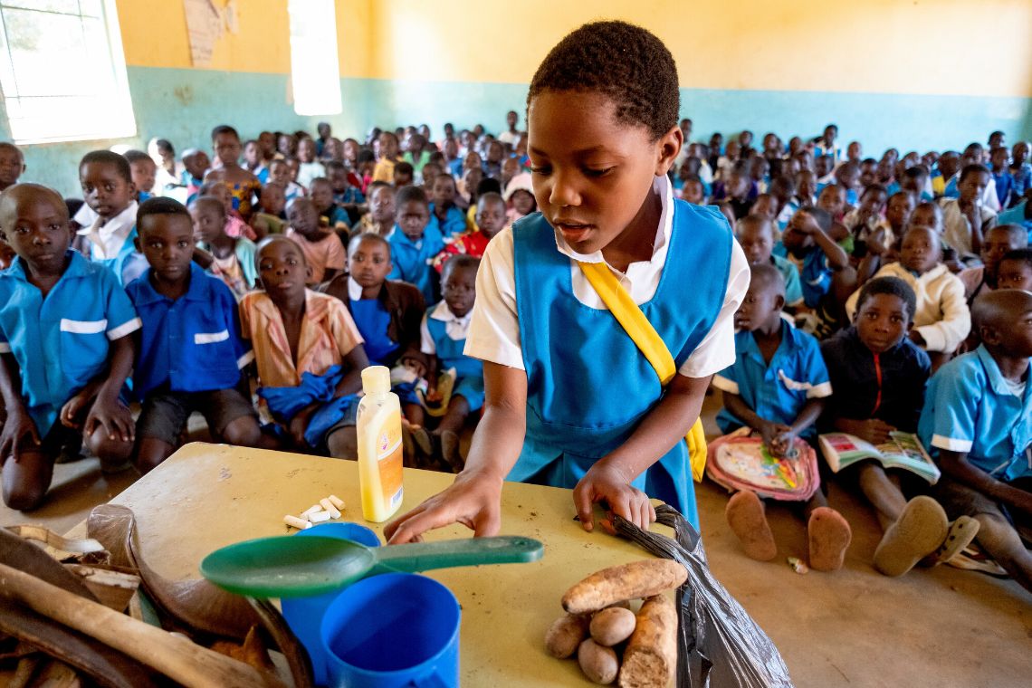 Ein Mädchen in Schuluniform steht an einem Pult in einem Klassenzimmer mit sehr vielen Kindern, die ihm auf dem Boden sitzen.